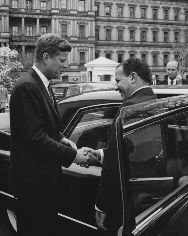 O Presidente dos Estados Unidos, John F. Kennedy, cumprimenta o Presidente do Brasil, João Goulart, em frente a um carro, na saída do Presidente Goulart da Casa Branca após sua reunião. O Executive Office Building é visível ao fundo. Entrada da ala oeste, Casa Branca, Washington, D.C.